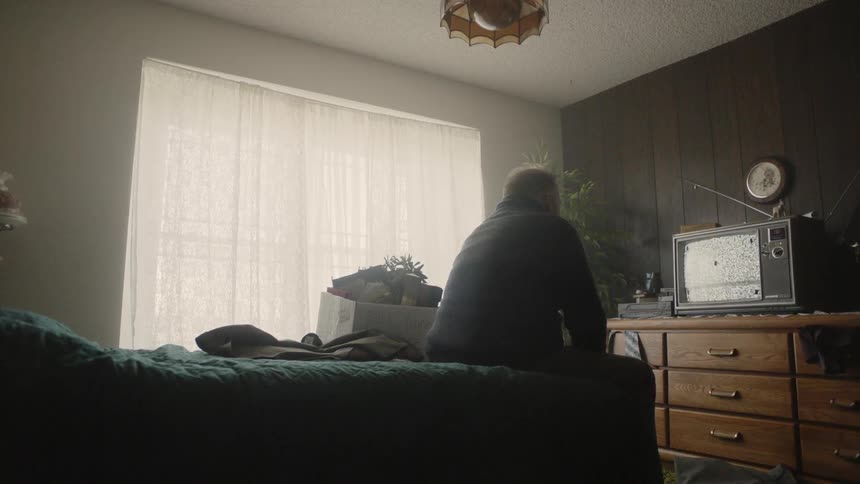 Man sits on the edge of a bed and stares at a television on the dresser