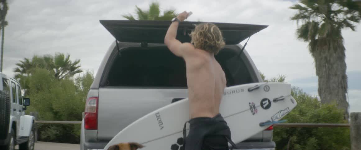 Man and a dog walk near a vehicle then the man takes a surfboard out from the back