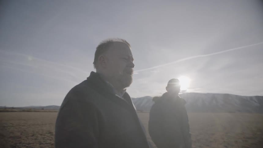 Two men walk in a rural field near mountains