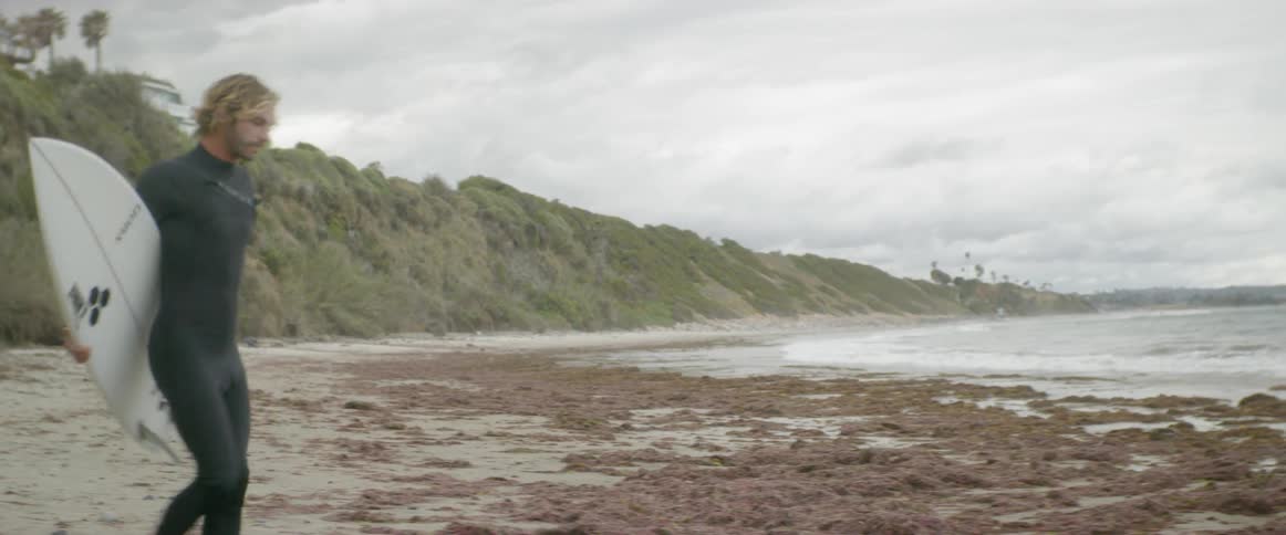 Dog runs ahead of a man walking with a surfboard on the beach