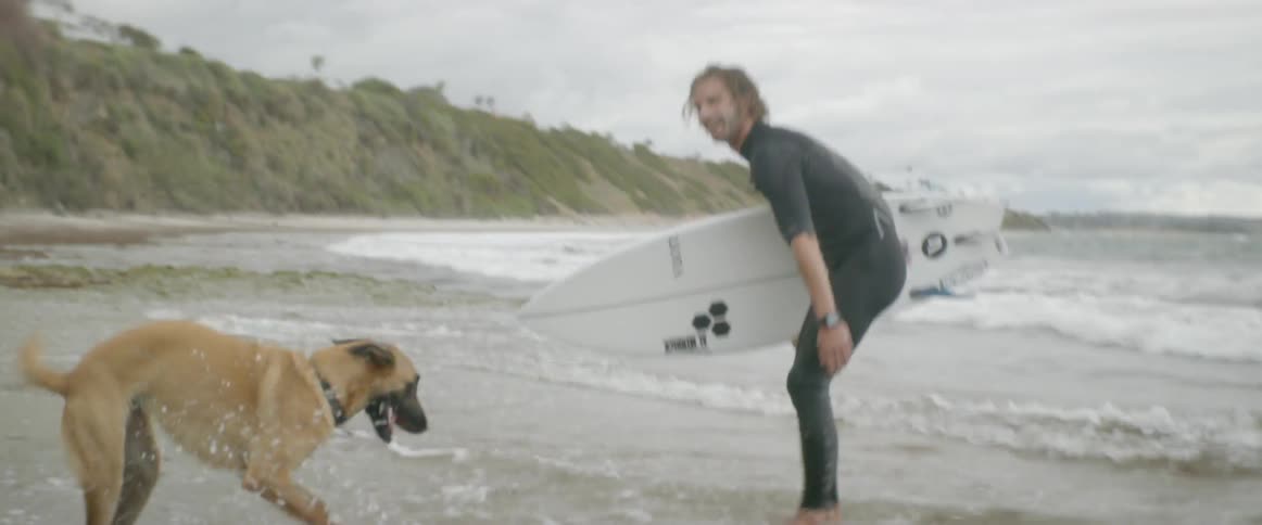 A man holding a surfboard plays with his dog on the beach