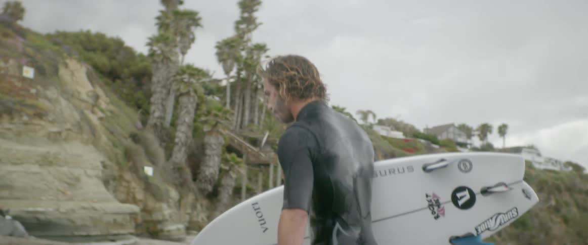 A man carrying a surfboard begins playing with his dog by the water