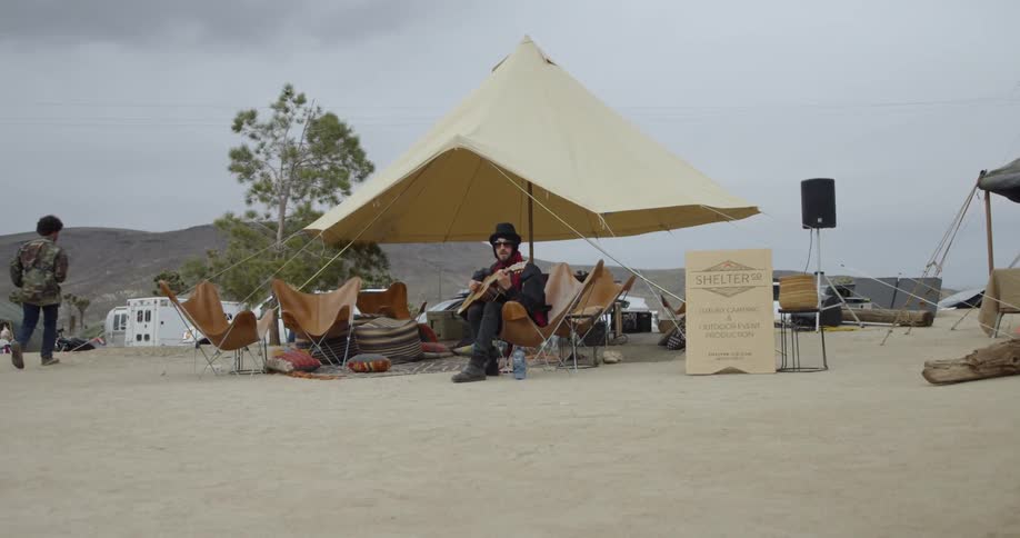 A man plays guitar while sitting in a chair under an umbrella