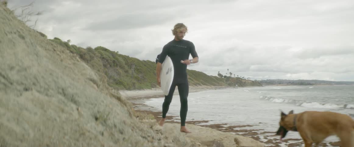 Man waxes his surfboard on the beach and walks away with his dog