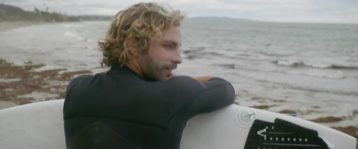 A dog comes up next to a man sitting on shore holding a surfboard