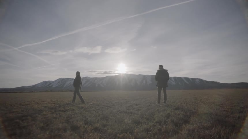 Two men walk in a rural field near mountains
