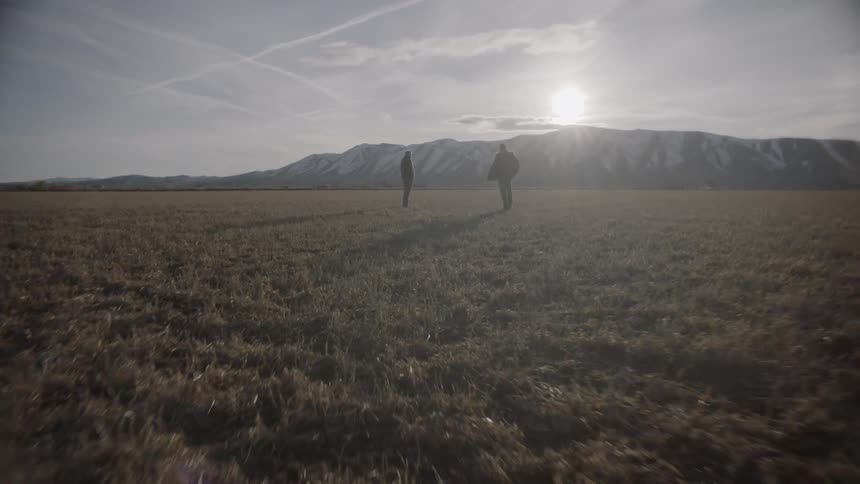 Two men stand in a rural field near mountains