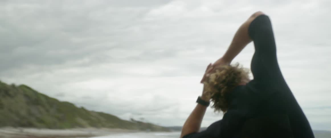 A man in a wet suit stretches on the beach and a dog walks by