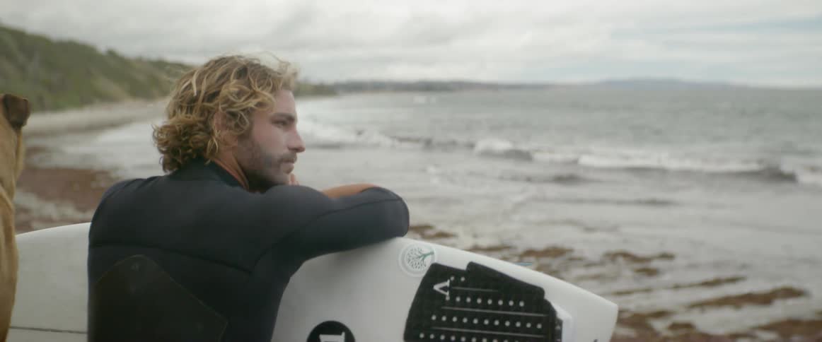 A man leans on his surfboard on the beach as the dog runs around