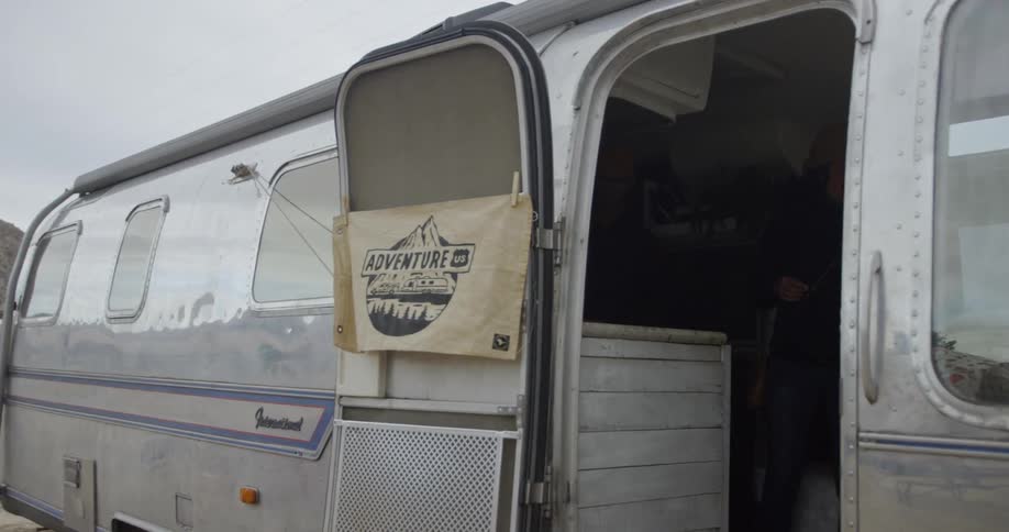 A sign is flapping in the wind on the door of a camper parked at a festival in daylight