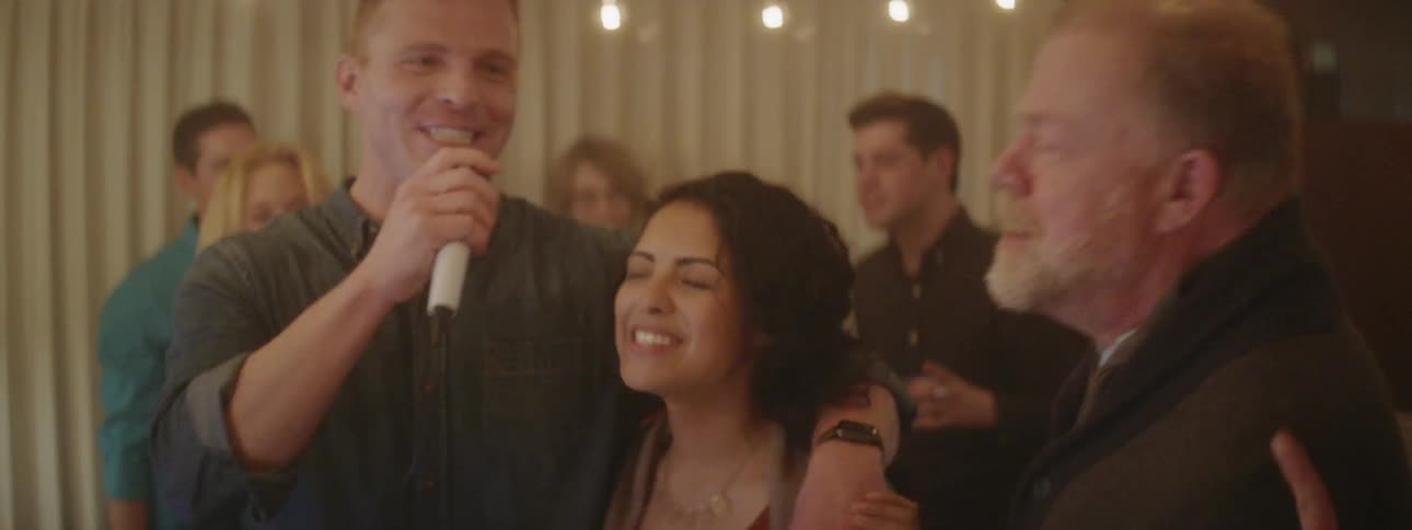 Man sings into a microphone as he has his arm around a woman