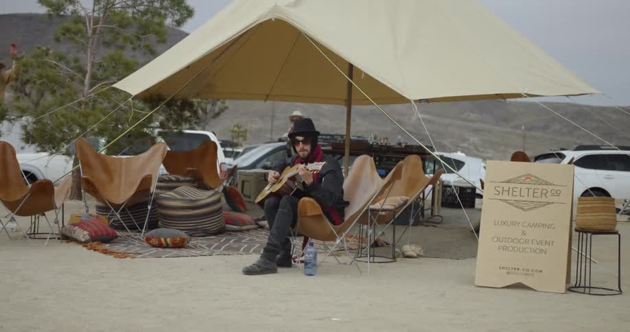 Man plays a guitar while sitting on a chair under a tent