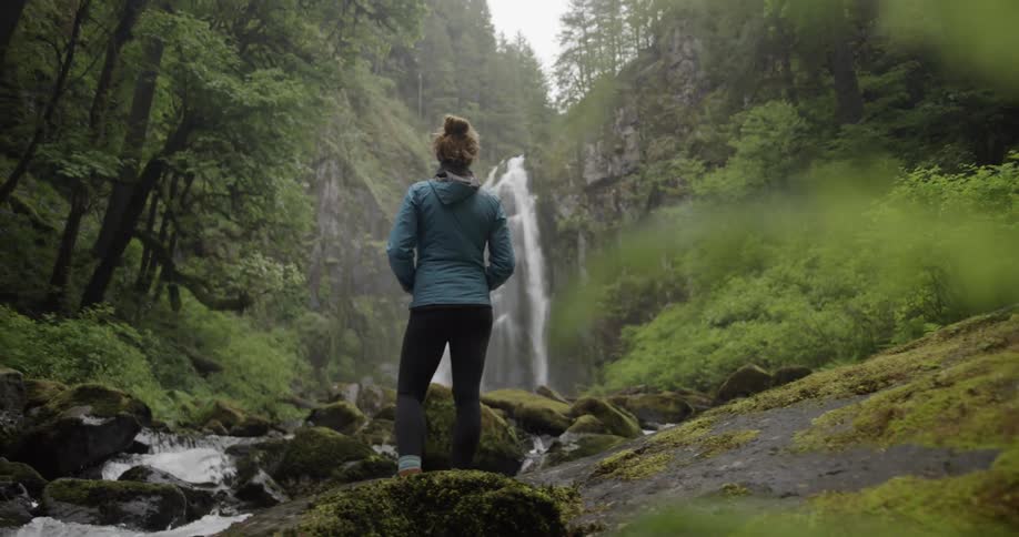 Dolly left shot of a woman in a green forest staring at a waterfall in a green forest
