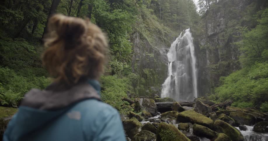 Woman stands in forest looking at a waterfall