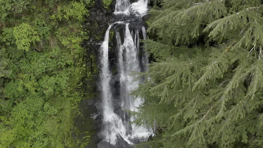 Aerial shot flying over a waterfall in a green forest flowing into a creek below