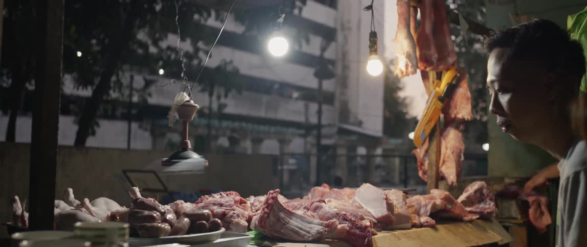 A fan hangs over meat on a butcher counter and turns as a man watches