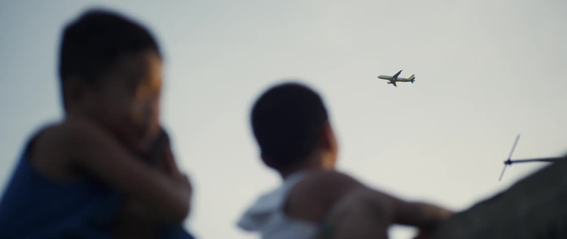 Three children stand outside looking up at an airplane