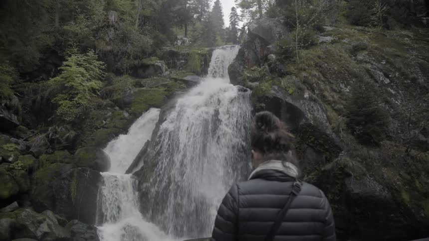 A woman is standing on a platform facing a waterfall in daylight