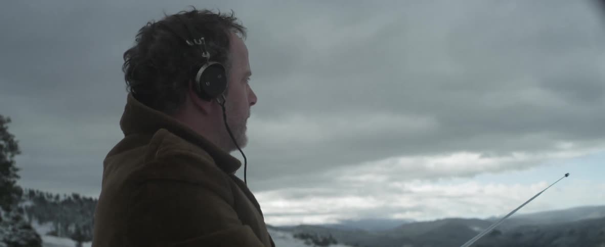 A man stands on top of the snowy mountain holding a device and listening with headphones