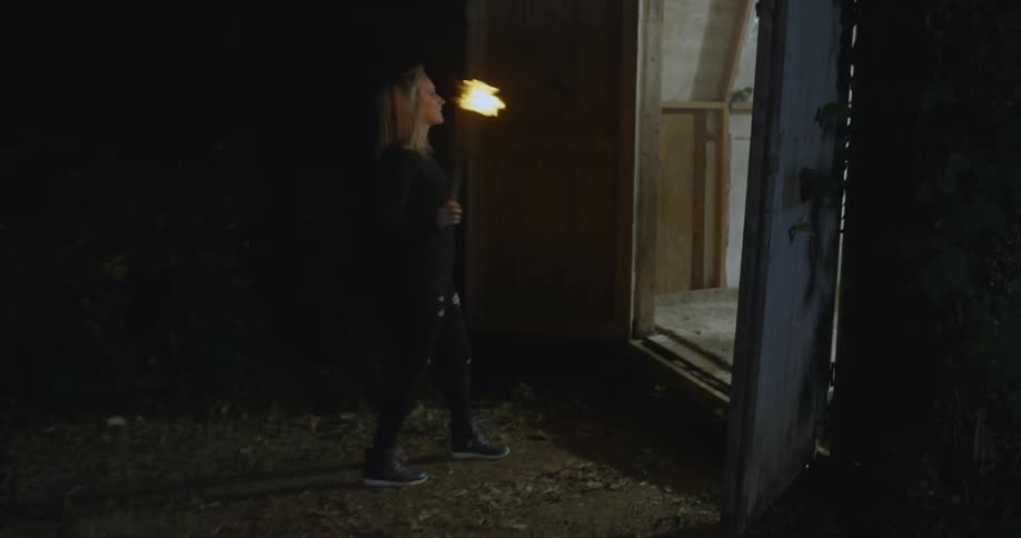 A woman is lighting a torch and then tossing it inside a building at night