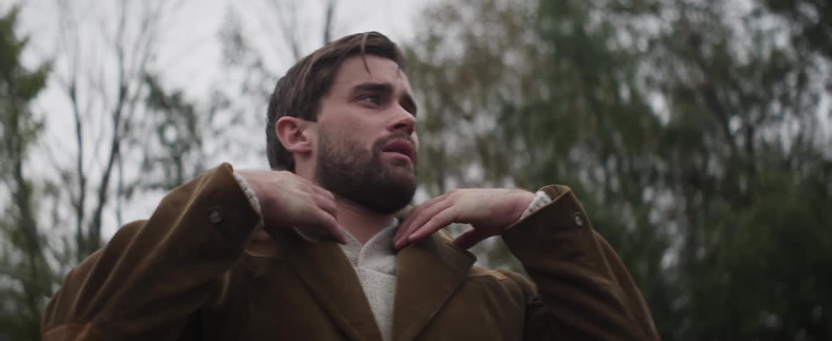 A man walking near trees and stopping to adjust his jacket