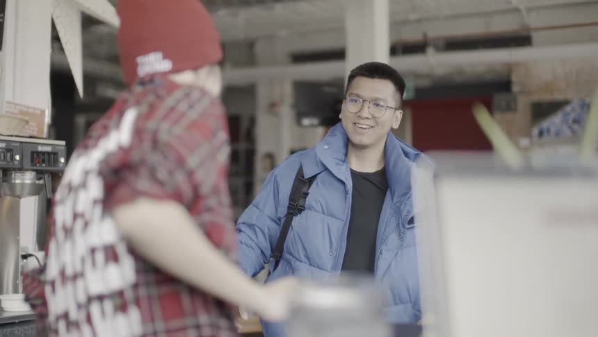 A man is entering an office space and greeting another man at a table