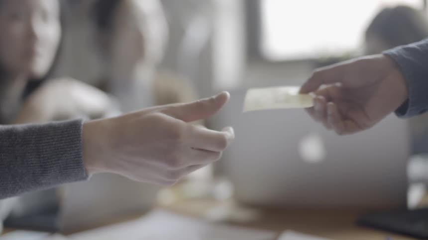 A person is handing a sticky-note to another person across a table inside in daylight