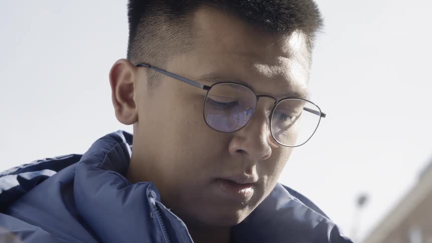 An asian man stands outside in a blue jacket