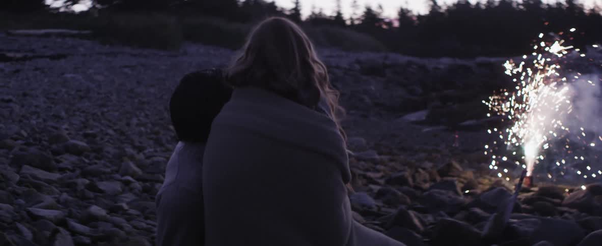 A couple sit on a rocky beach watching a small display of sparks at dusk