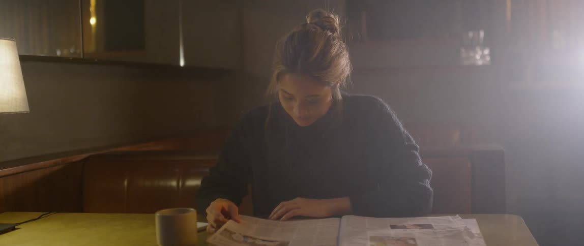A woman sits with a drink and a newspaper