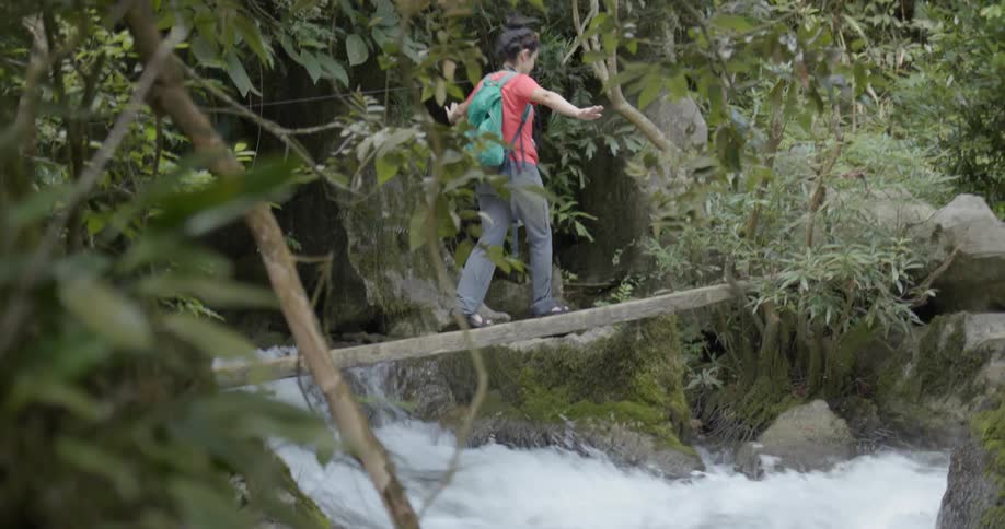 A woman is balancing on a wood plank to cross a rushing river during the day