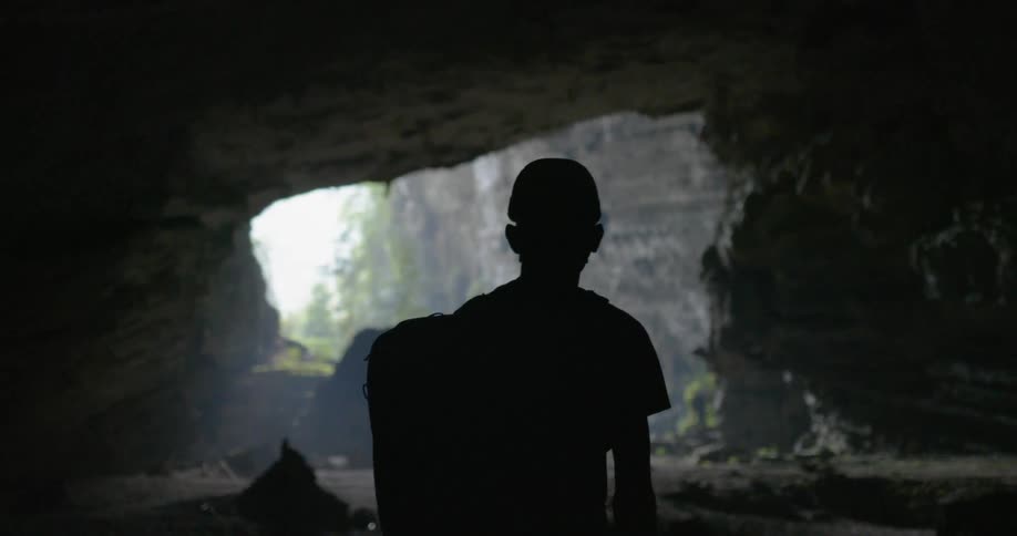 A person wearing a backpack stands in a cavern looking out