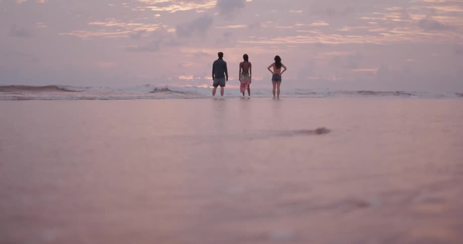 Man and two woman are standing on ocean beach at sunset