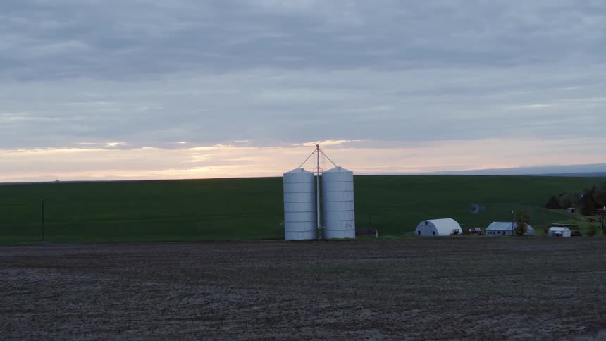 Flying toward lush cropland as silos stand on a farmstead at sunset