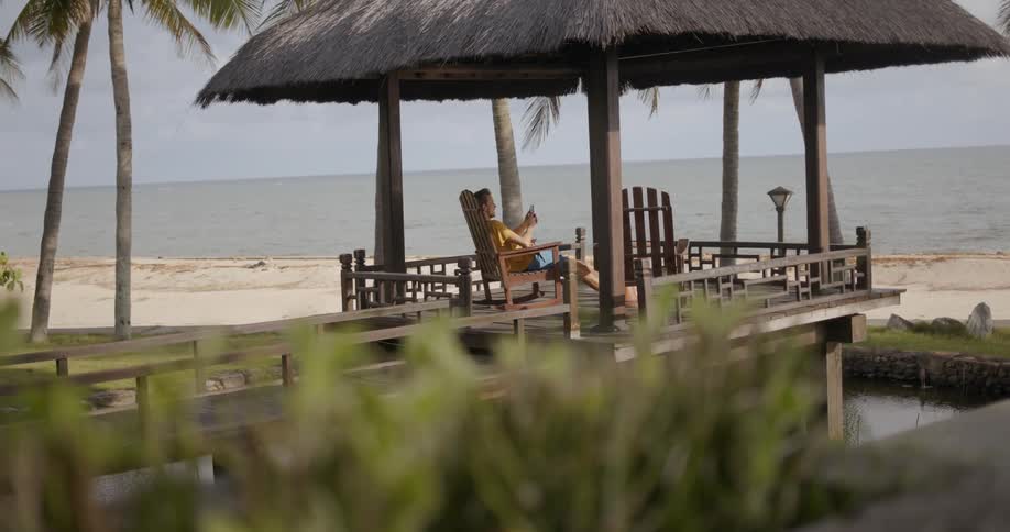 A man sits on a chair under a pergola on the beach on a slightly cloudy day