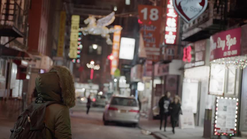 A person wearing a parka with a hood up looks around at the buildings and signs