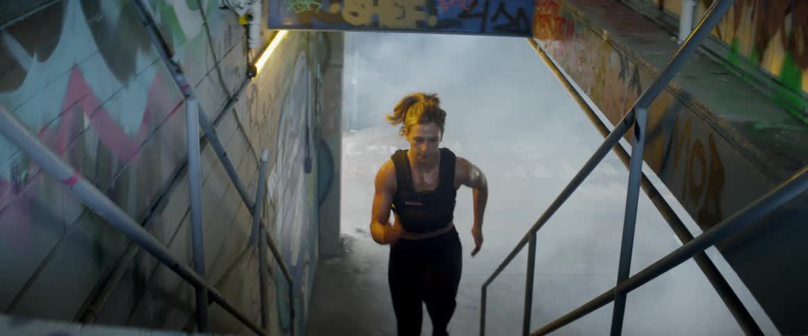 A woman runs through a smoky area up a staircase into an industrial building with graffiti on the walls