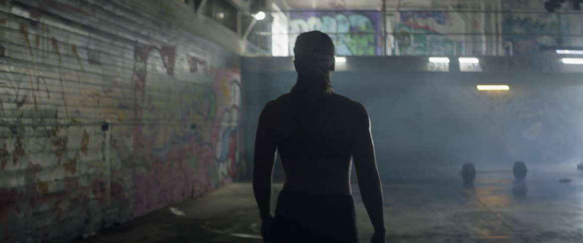 Woman walks towards barbells in dimly lit gym