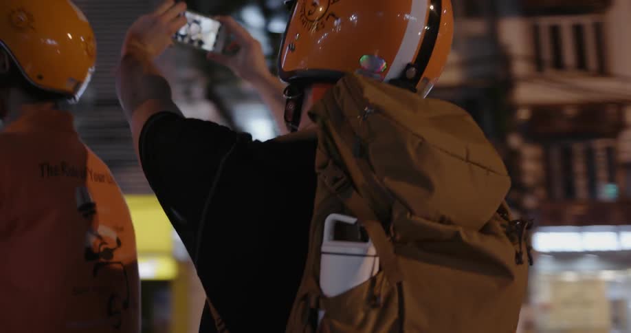 Two people ride on a motorbike through a city at night and one takes a picture