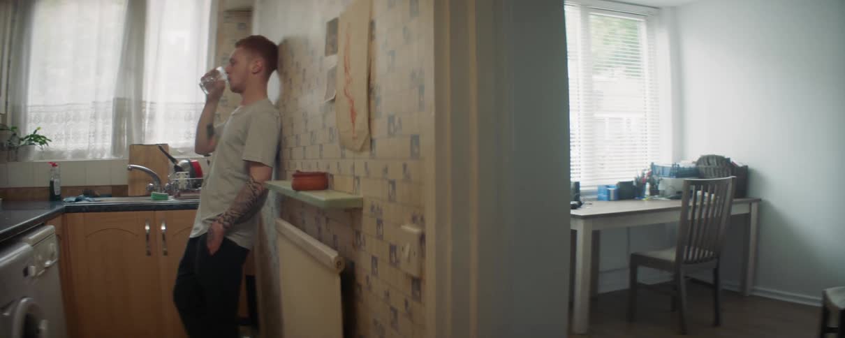 A man is leaning against the wall drinking in the kitchen