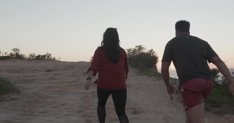Man and woman are walking up hill overlooking ocean at sunset