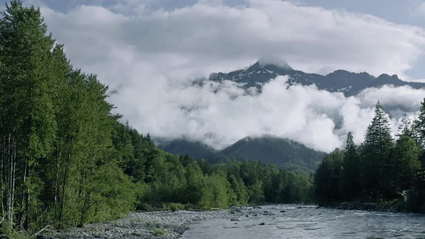 Clouds pass near the tall mountains above the river - 4K Aerial