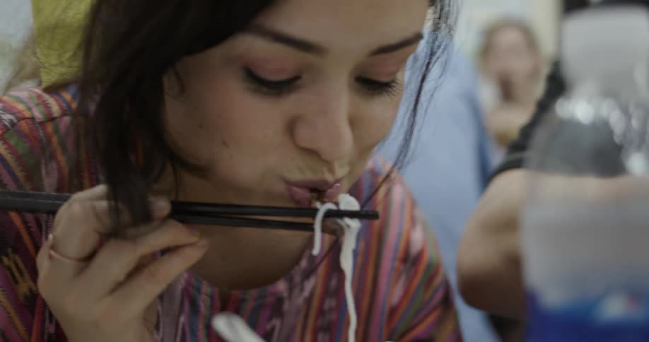 A woman sits at a table smiling and eating a dish of noodles