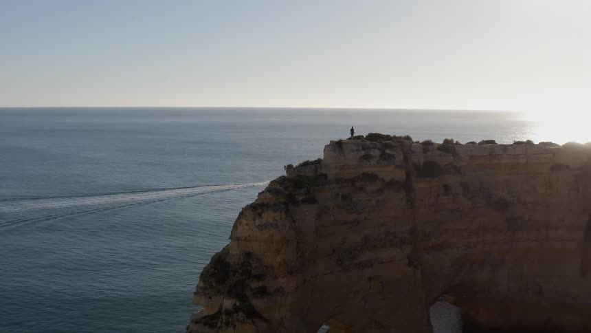 Person standing on huge rock cliff overlooking ocean at sunset