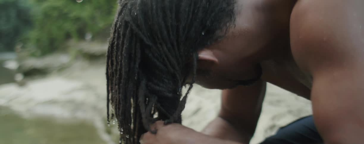 Man is wringing out water from dreadlocks by river