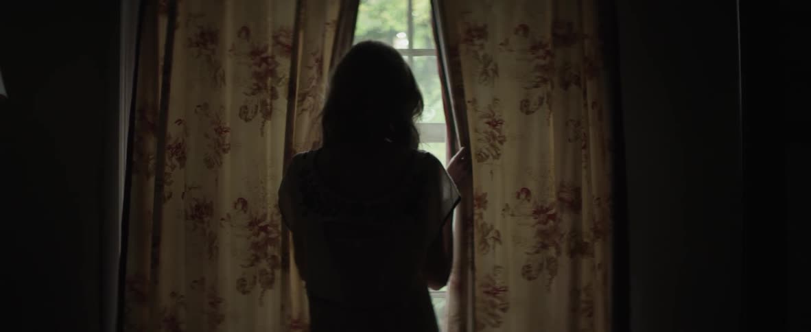 Woman walks over to a window and peaks through the blinds