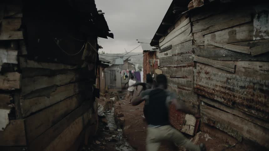 A gang of boys are chasing another boy through a slum on a cloudy day