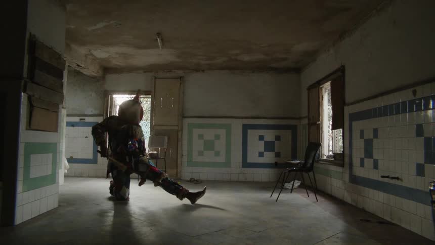 Person wearing a costume dances in an empty room waving two sticks