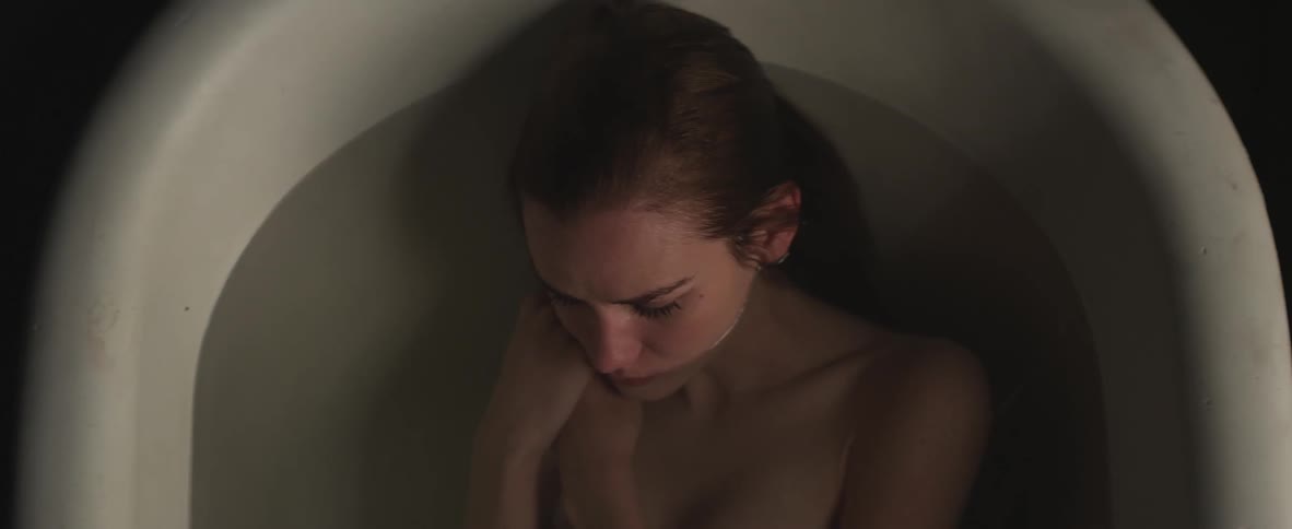 A young woman lays naked in a filled bathtub while thinking
