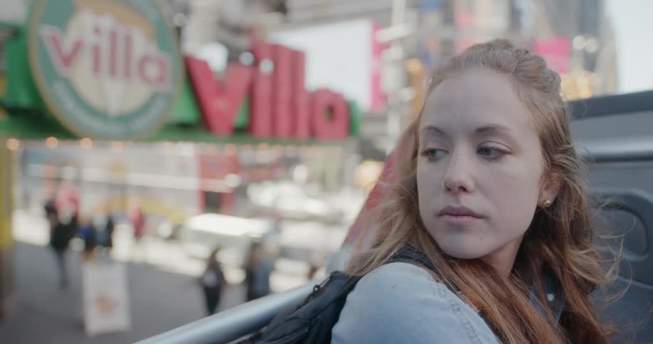 A woman sits outdoors above people walking on a street and flashing signs